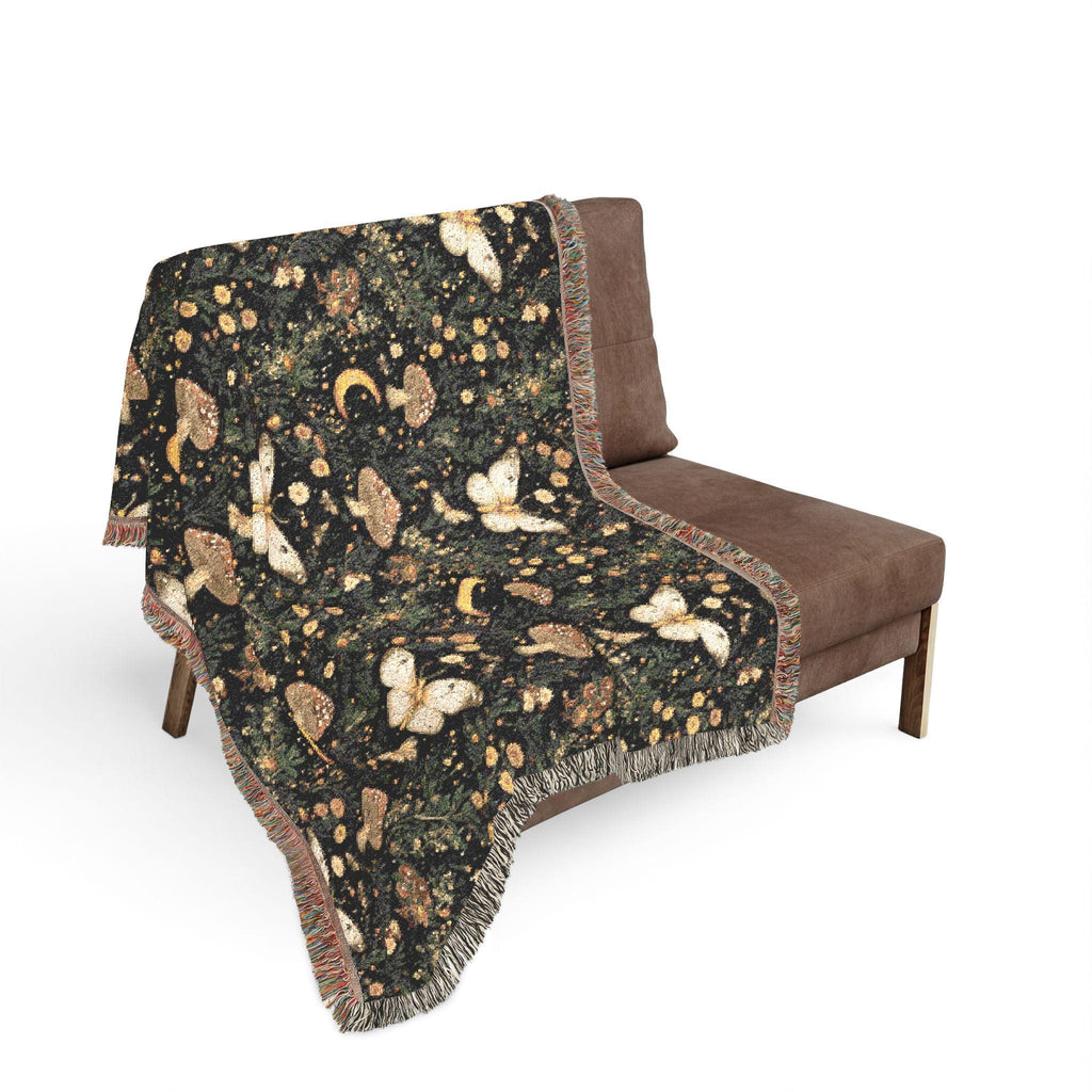 A woven throw blanket with a celestial, nature-inspired pattern featuring butterflies, moons, and mushrooms on a dark background, draped over a brown armchair against a white background.