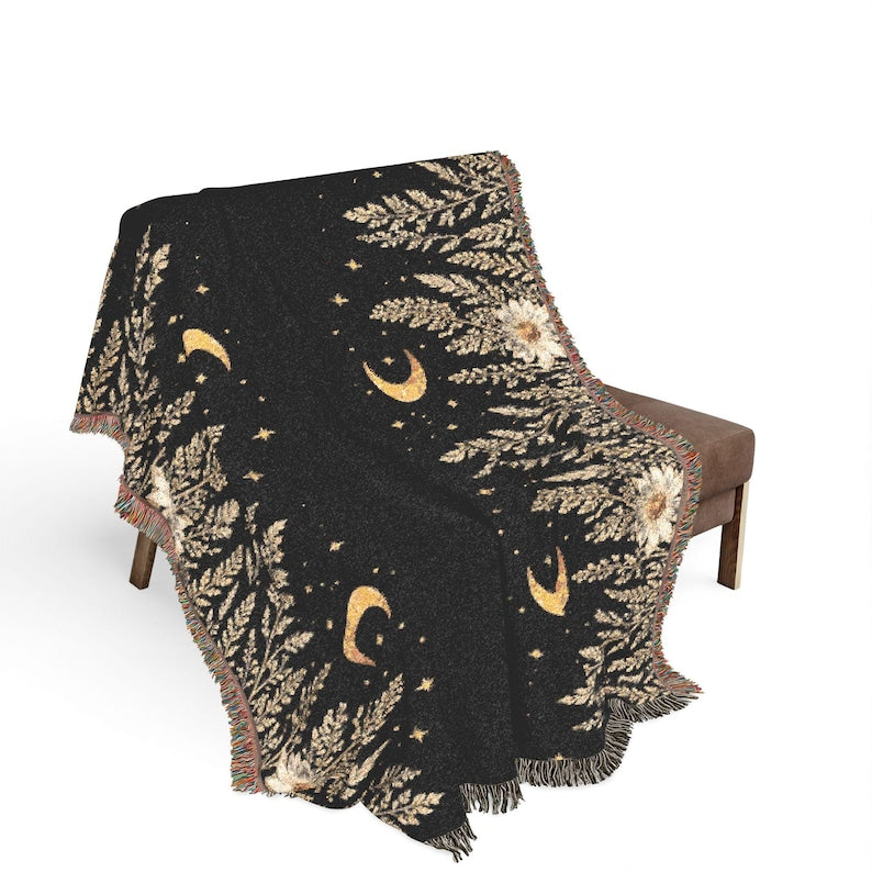 Black woven throw blanket with gold crescent moons, stars and botanical floral details, displayed over a chair