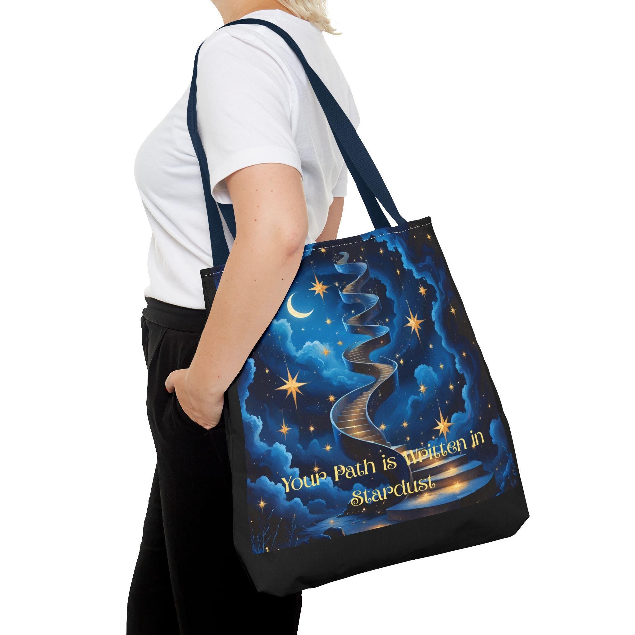 Woman carrying Cosmic Stardust Tote Bag at side — deep blue night sky design with gold stars, crescent moon, stardust path, and script quote, navy handle