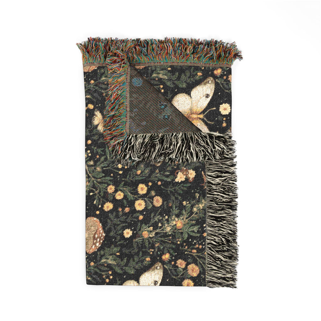 A rectangular woven throw blanket with fringed edges, featuring a dark forest pattern with butterflies, flowers, and foliage. One corner is folded to reveal the reverse side with a textured geometric pattern and blue accents.