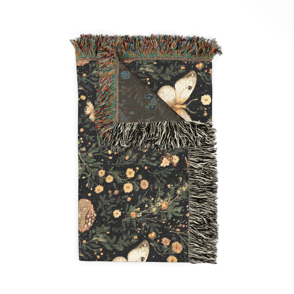 A rectangular woven throw blanket with fringed edges, featuring a dark forest pattern with butterflies, flowers, and foliage. One corner is folded to reveal the reverse side with a textured geometric pattern and blue accents.
